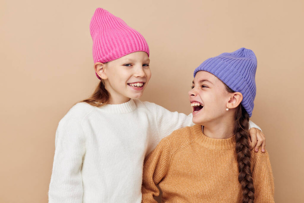 two little girls in sweaters and hats hug friendship beige background - Фото, изображение