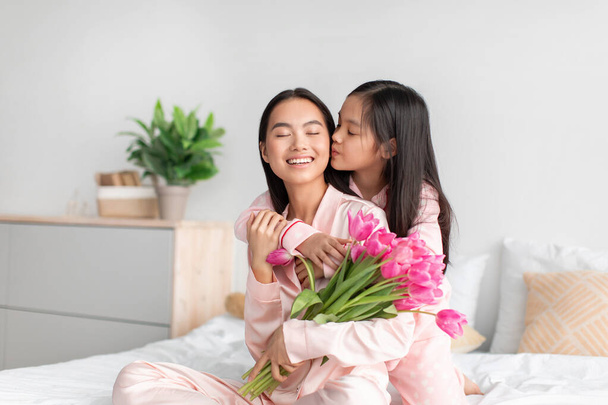 Gülümseyen mutlu Asyalı genç kız yatak odasında bir buket lale ile milenyum kadınını öpüyor ve sarılıyor. - Fotoğraf, Görsel