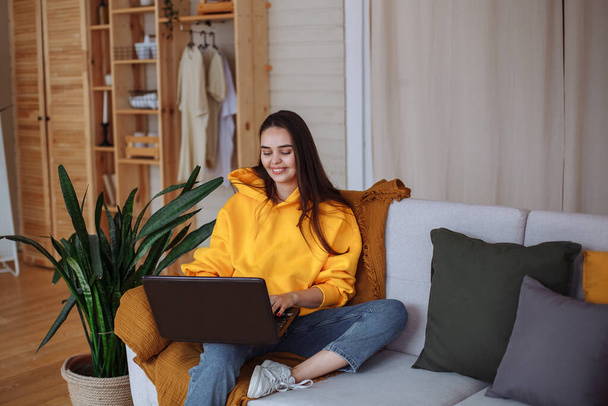 A smiling young brunette woman with long hair in a yellow sweatshirt, blue jeans and white sneakers sits at home on a gray sofa with a laptop. There is a mustard blanket on the sofa. Home green plants stand nearby - Foto, afbeelding