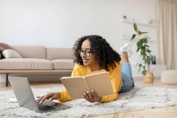 Jonge zwarte vrouw lezen boek in de buurt van laptop, klaar voor examen of werken aan project, liggend op de vloer thuis - Foto, afbeelding