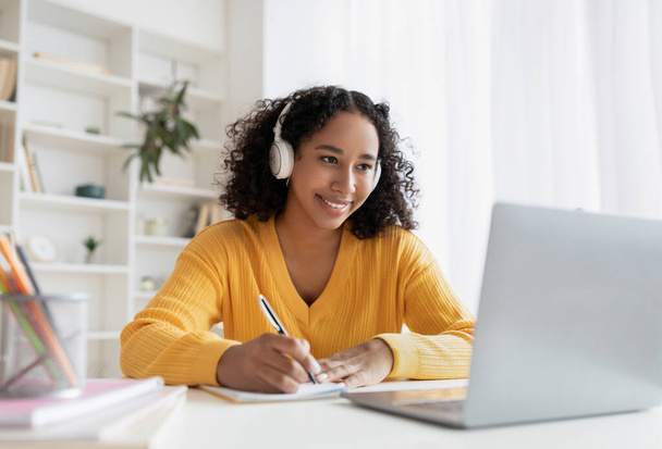 Happy African American woman in headphones having online meeting on laptop, writing down info during distance lesson - Foto, Imagen