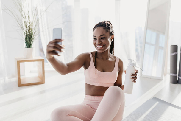 Affascinante giovane donna afroamericana con bottiglia d'acqua che prende selfie durante l'allenamento a casa - Foto, immagini