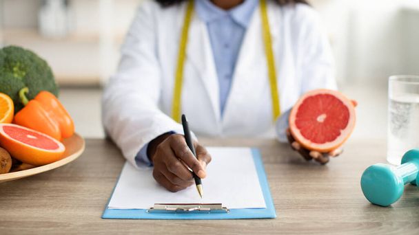 African american female dietologist holding grapefruit and writing healthy eating plan or weight loss diet for patient - Foto, Imagem