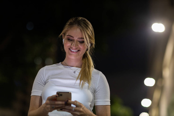 Young woman using a smartphone at night time with city view landscape in the background. High quality photo. photo. Mobile phone, technology, urban concept.  - Zdjęcie, obraz