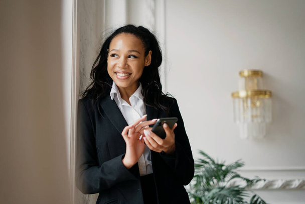 It works by printing a message to the client on the phone in a coworking space. Confident business woman smiling afro appearance sitting in the office in a formal suit - Fotografie, Obrázek