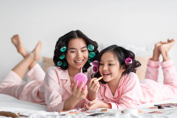 Χαρούμενη χιλιετή γιαπωνέζα κυρία με μπικουτί κρατά καθρέφτη για έφηβο κορίτσι, απλώνει κραγιόν στα χείλη, στο κρεβάτι με καλλυντικά - Φωτογραφία, εικόνα
