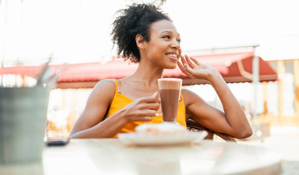  He has breakfast, drinks cocoa, and has breakfast. A woman of African American appearance smiles sitting in a summer restaurant. - Foto, imagen
