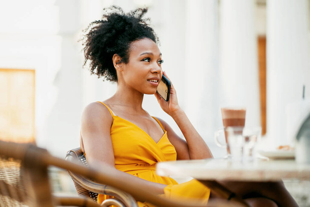 A freelance blogger is talking on the phone with a friend and smiling. Female African American appearance, lunch break. Uses a new high-quality 5G cellular connection. - Foto, afbeelding