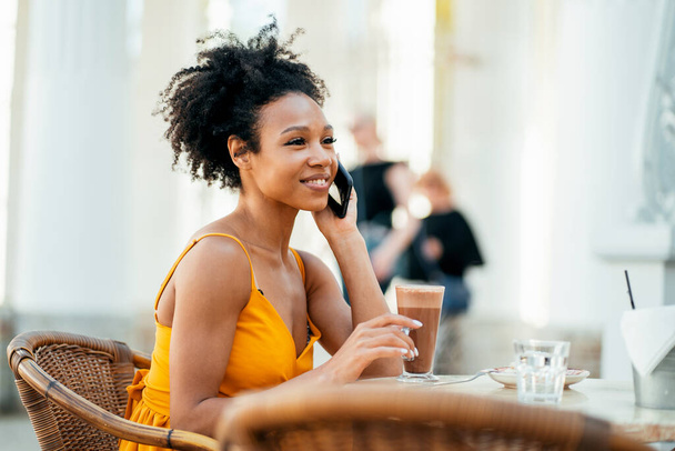 Drinks cocoa and has breakfast. Makes an online order in the online store on the website. A woman of Afro appearance smiles sitting in a summer restaurant. - Photo, Image