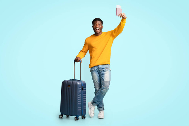 Χαρούμενος Αφροαμερικάνος με βαλίτσα που δείχνει αεροπορικά εισιτήρια - Φωτογραφία, εικόνα