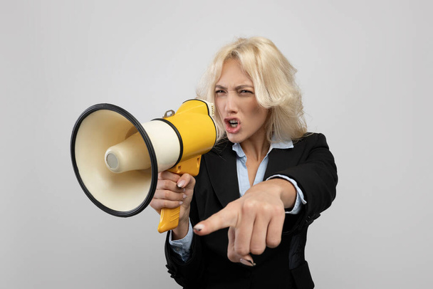 Agitated businesswoman in formalwear shouting into megaphone and pointing finger, posing on grey background - 写真・画像
