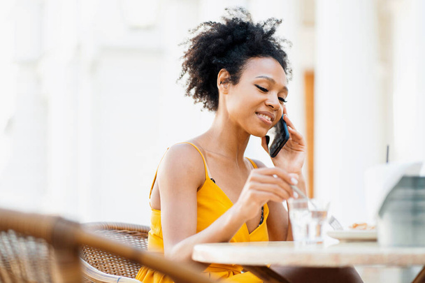 Manager online work remotely. Freelance Afro ethnic appearance pays for purchases on the phone in a summer cafe. A female blogger responds to clients in a chat. - Foto, afbeelding
