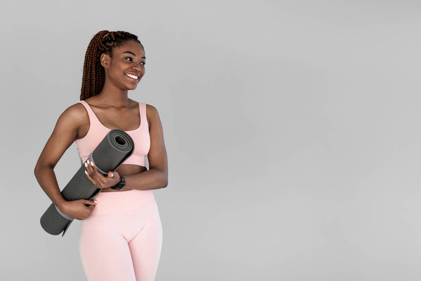 Młoda czarna dama w odzieży sportowej trzymająca matę fitness, uśmiechnięta i patrząca na puste miejsce na szarym tle studia - Zdjęcie, obraz