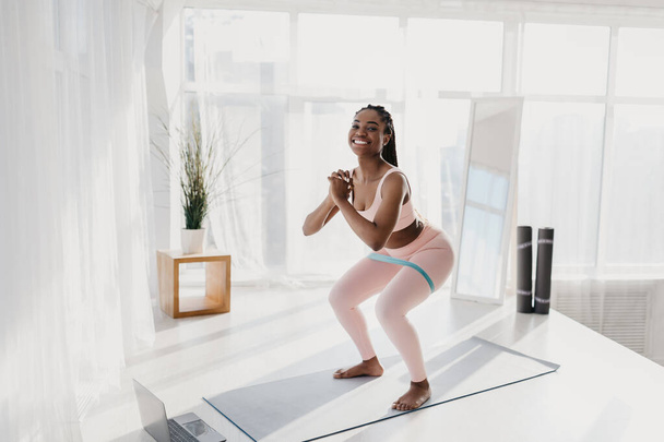 Full length of fit young African American woman squatting with elastic band on mat, exercising online at home, copy space - Foto, imagen