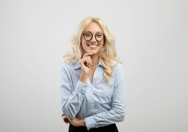 Женская бизнес-карьера. Позитивная молодая деловая женщина касается подбородка и улыбается в камеру, стоя на светлом фоне - Фото, изображение