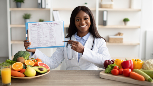 Concepto nutricional correcto. Nutricionista negro feliz mostrando plan de dieta semanal, sentado en el escritorio con frutas y verduras - Foto, Imagen