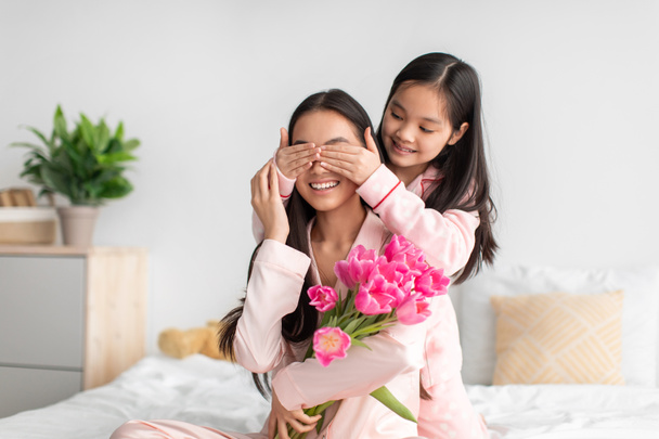 Lächelnde junge Chinesin schließt Augen vor Millennial-Weibchen mit Tulpenstrauß im Schlafzimmer - Foto, Bild