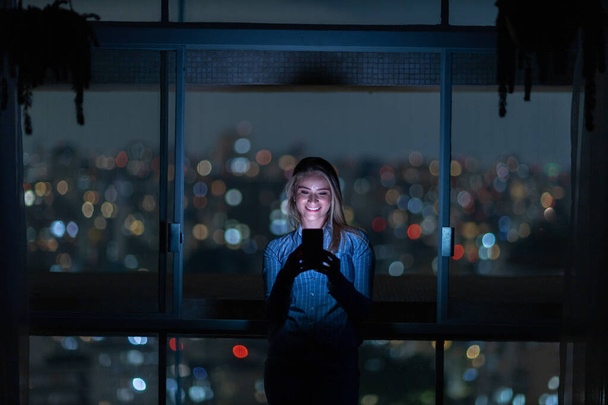 Young woman using a smartphone at night time with city view landscape in the background. High quality photo. Mobile phone, technology, concept. High quality photo - Fotó, kép