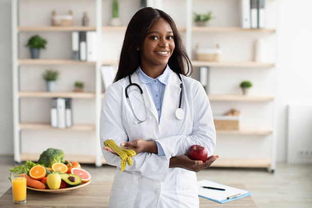 Портрет веселой молодой черной женщины-диетолога с яблоком и лентой, рекомендующей фрукты и овощи, бесплатное место - Фото, изображение