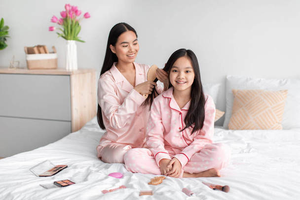 Souriant jeune asiatique femelle peignage longs cheveux lisses de l'adolescente, assis sur le lit avec des cosmétiques dans la chambre à coucher intérieur - Photo, image