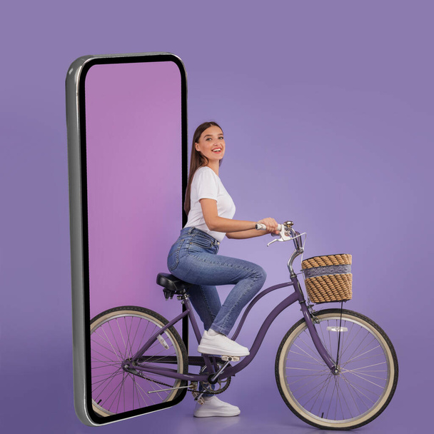 Happy Lady andar de bicicleta perto de grande celular sobre fundo roxo - Foto, Imagem