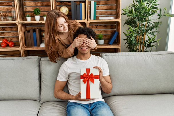 Young couple smiling happy sitting on the sofa surprise with anniversary gift at home. - Photo, Image
