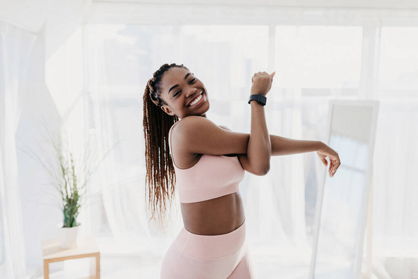 Portrait of attractive fit African American lady in sportswear exercising at home, stretching her arm and smiling at camera - Zdjęcie, obraz