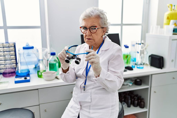 Oudere vrouw met grijs haar werkt in het laboratorium met behulp van vergrootglazen depressief en zorgen te maken voor nood, huilen boos en bang. trieste uitdrukking.  - Foto, afbeelding