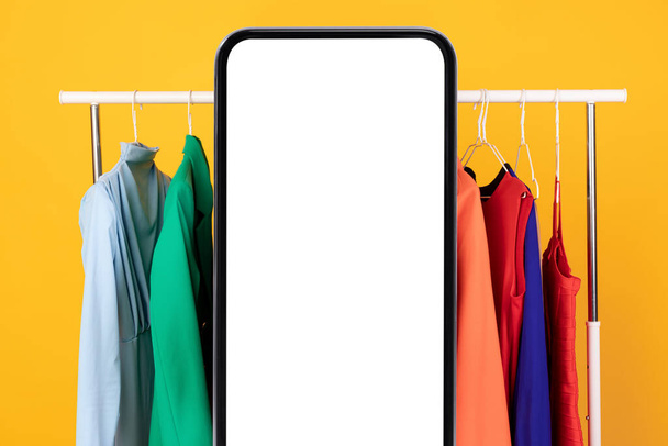 Gran Smartphone pantalla en blanco de pie cerca del carril de la ropa, fondo amarillo - Foto, Imagen