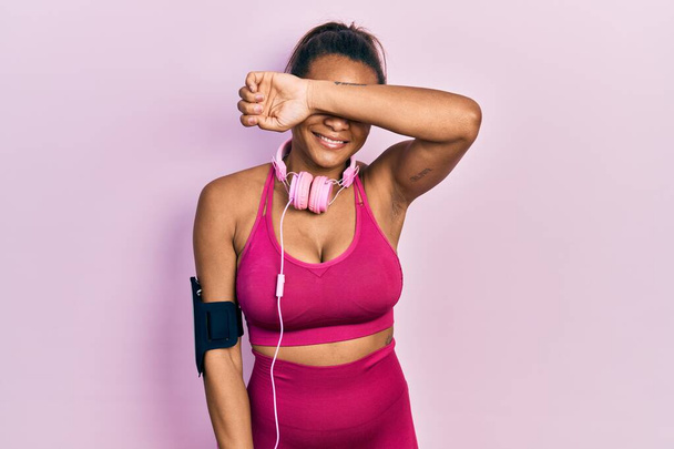 Young african american girl wearing gym clothes and using headphones covering eyes with arm smiling cheerful and funny. blind concept.  - Foto, immagini