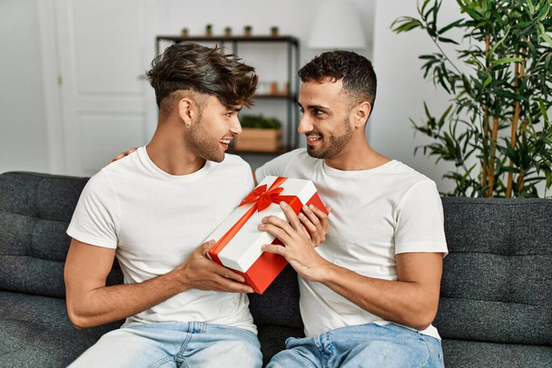 Dos hombres hispanos pareja sonriendo confiada sorpresa con regalo en casa - Foto, imagen