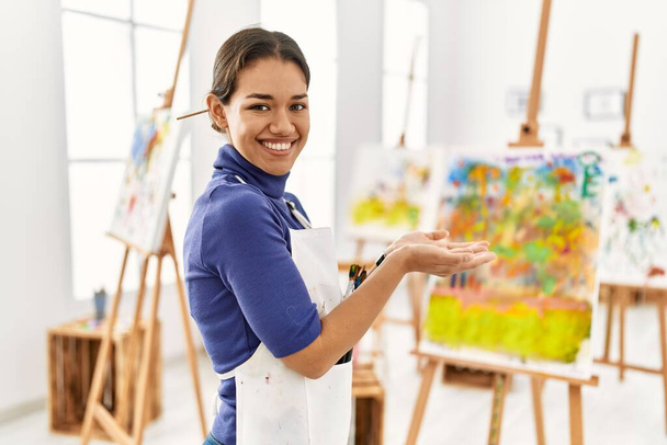 Jeune femme brune au studio d'art pointant de côté avec les mains ouvertes paumes montrant l'espace de copie, présentant la publicité souriant excité heureux  - Photo, image