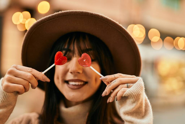 Brunette woman wearing winter hat being funny holding lollipops covering eyes outdoors at the city - Foto, Bild