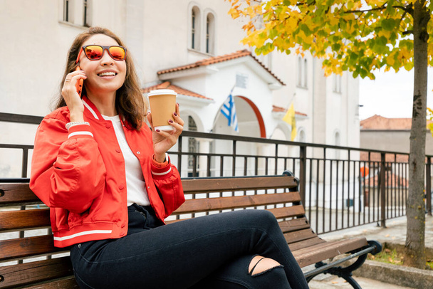 Egy mosolygó lány telefonál és forró kávét iszik egy padon a parkban. - Fotó, kép