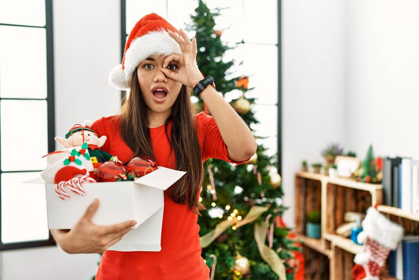 Jeune femme brune debout près du sapin de Noël tenant la décoration faisant un geste correct choqué par le visage surpris, les yeux regardant à travers les doigts. expression incrédule.  - Photo, image