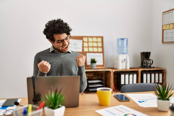 Young hispanic man wearing business style sitting on desk at office very happy and excited doing winner gesture with arms raised, smiling and screaming for success. celebration concept.  - Photo, Image