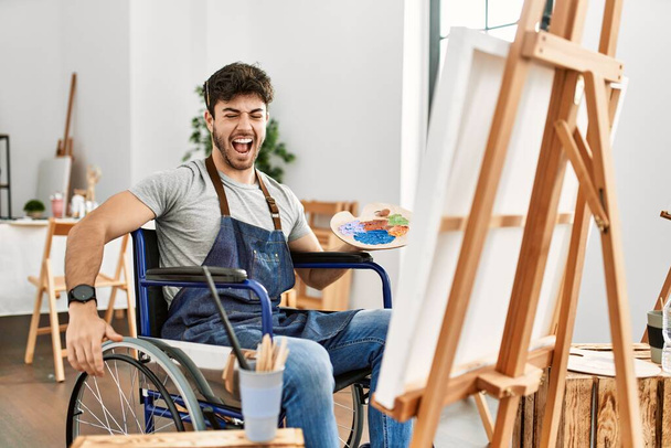 Joven hombre hispano sentado en la pintura en silla de ruedas en el estudio de arte enojado y loco gritando frustrado y furioso, gritando con ira. rabia y concepto agresivo.  - Foto, Imagen