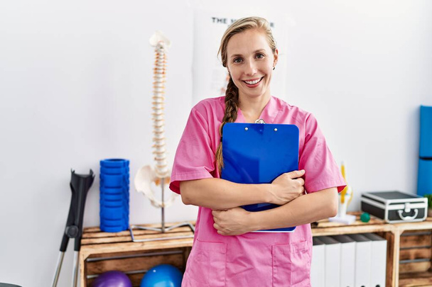 Jonge blanke vrouw draagt fysiotherapeut uniform met checklist staande in fysiotherapie kliniek - Foto, afbeelding