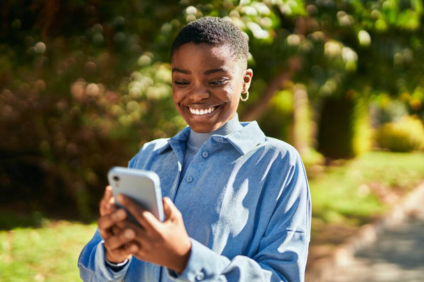 Νεαρή Αφροαμερικανή γυναίκα χαμογελά ευτυχισμένη χρησιμοποιώντας smartphone στην πόλη - Φωτογραφία, εικόνα