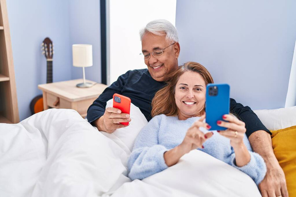 Mężczyzna i kobieta w średnim wieku para za pomocą smartfona leżącego na łóżku w sypialni - Zdjęcie, obraz