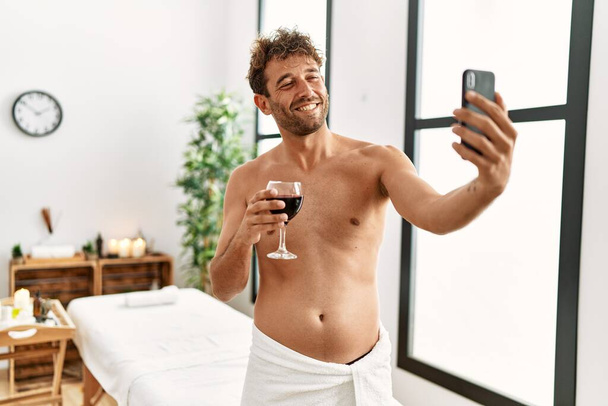 Молодий іспаномовний чоловік, що п'є вино, робить селфі біля смартфона в центрі краси
 - Фото, зображення