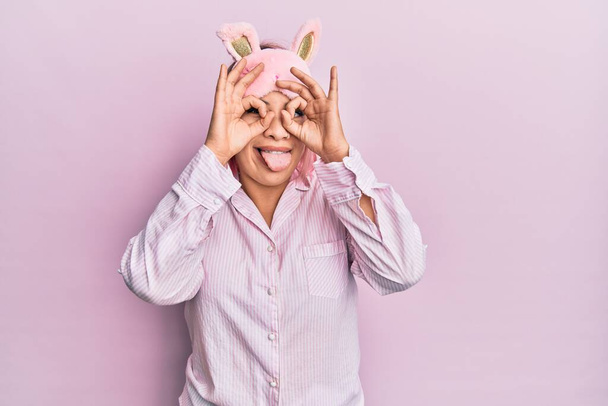 Латиноамериканка с розовыми волосами в маске для сна и пижаме делает нормальный жест, как бинокль высовывает язык, глаза смотрят сквозь пальцы. сумасшедшее выражение.  - Фото, изображение