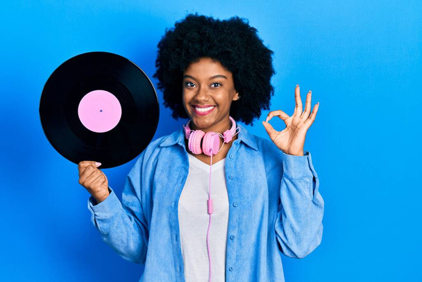 Молодая африканская американка в наушниках с виниловым диском делает хорошо знак с пальцами, улыбаясь дружелюбный жест отличный символ  - Фото, изображение
