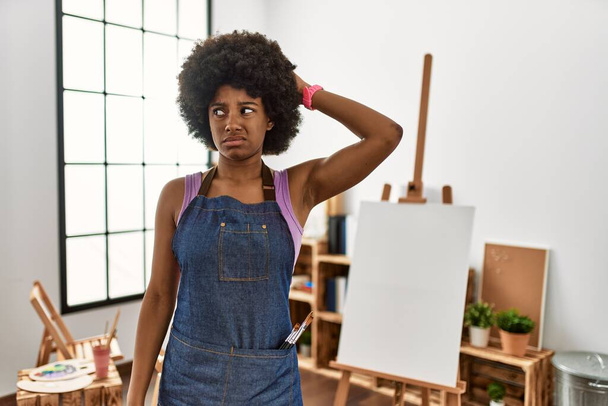 Молодая африканская американка с афроволосами в художественной студии сбивает с толку и задается вопросом. неуверенный в сомнении, думающий с рукой на голове. застенчивое понятие.  - Фото, изображение