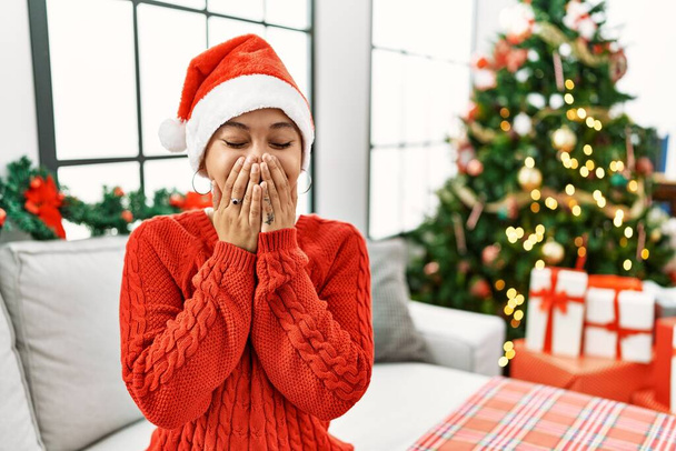 Fiatal spanyol nő rövid haj visel karácsonyi kalap ül a kanapén nevetve és zavarban vihog elfedi a száját kézzel, pletyka és botrány koncepció  - Fotó, kép