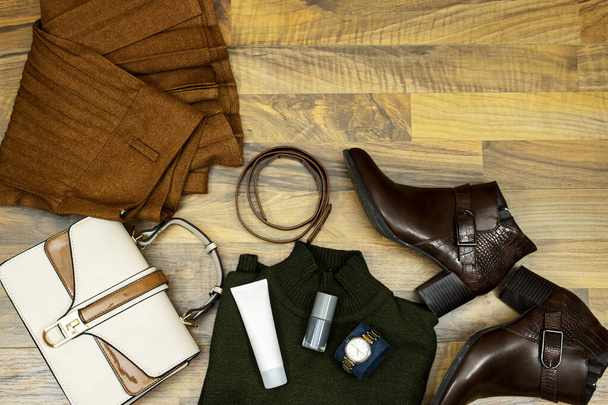 Μια σειρά από μοντέρνα ρούχα και αξεσουάρ. πράσινο πουλόβερ και κυματοειδές φούστα, τσάντα, μπότες αστράγαλο και καφέ δερμάτινη ζώνη και σωλήνα κρέμα προσώπου, βερνίκι νυχιών σε ξύλινο τραπέζι, επίπεδη lay. Να είστε στην τάση - Φωτογραφία, εικόνα