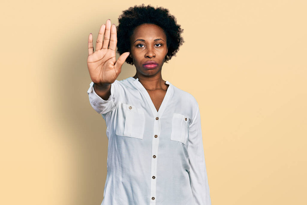 Femme afro-américaine aux cheveux afro portant un t-shirt blanc décontracté qui arrête de chanter avec la paume de la main. expression d'avertissement avec geste négatif et sérieux sur le visage.  - Photo, image