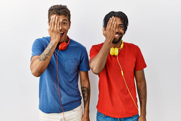 Fiatal spanyol testvérek állnak elszigetelt háttér visel fejhallgató, amely az egyik szemét kézzel, magabiztos mosoly az arcon és meglepetés érzelem.  - Fotó, kép