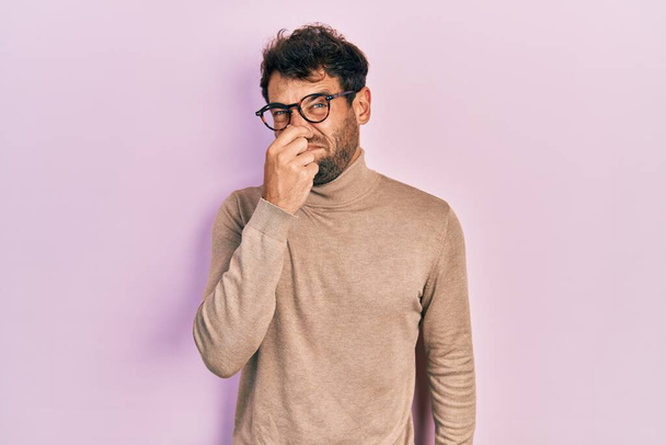 Handsome man with beard wearing turtleneck sweater and glasses smelling something stinky and disgusting, intolerable smell, holding breath with fingers on nose. bad smell  - Photo, Image