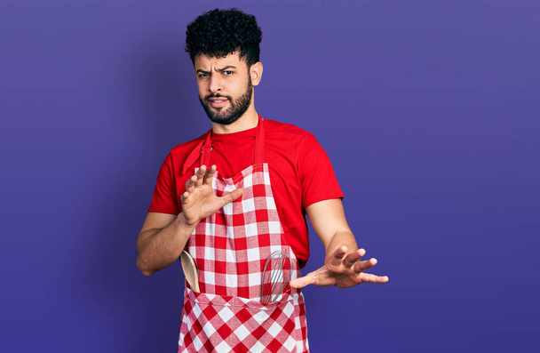 Mladý arabský muž s vousy v kuchařské zástěře znechucený výraz, nespokojený a ustrašený dělá znechucený obličej, protože averzní reakce. s rukama zvednutýma  - Fotografie, Obrázek